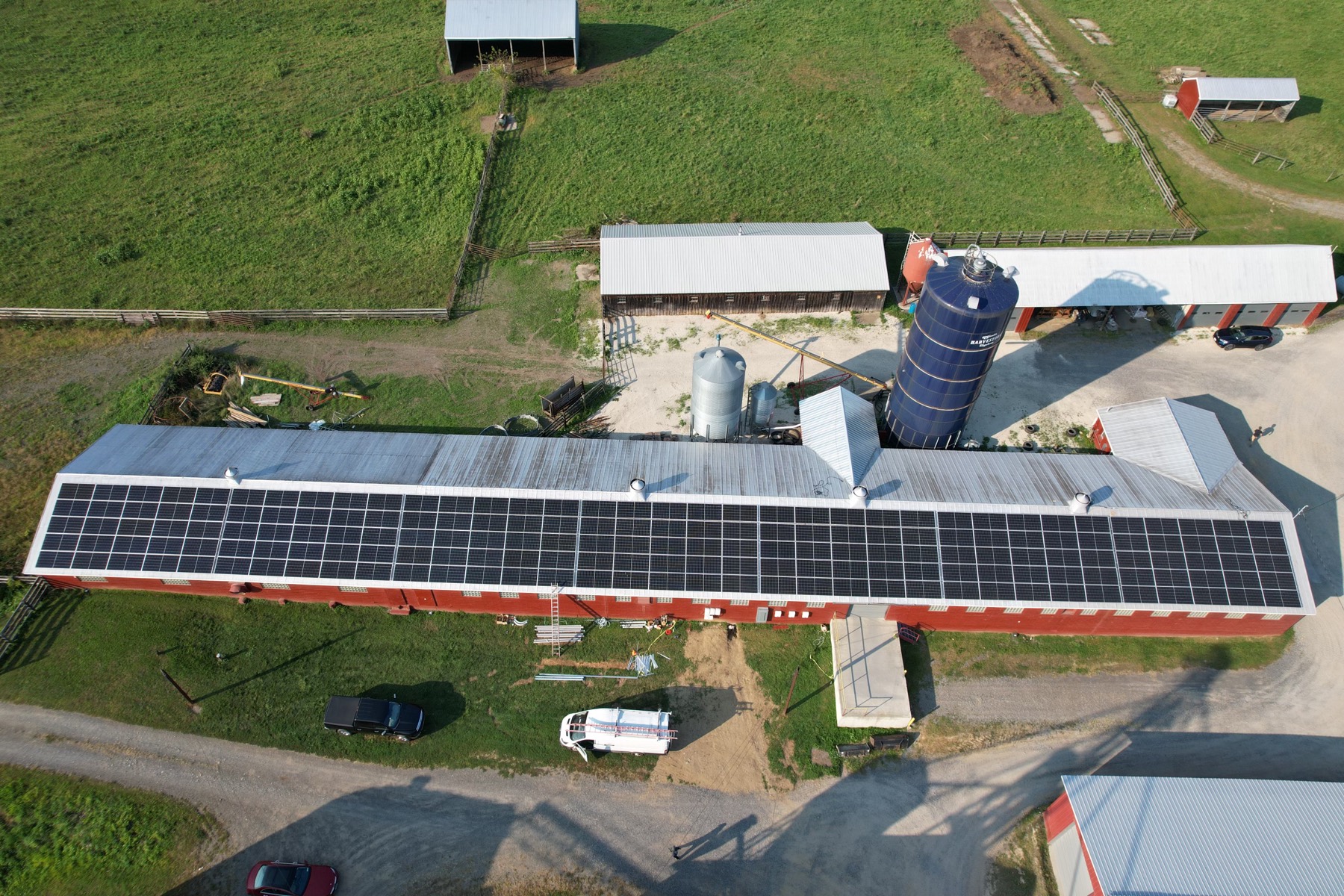 Aerial view of a large commercial solar array covering the roof of a long red farm barn, with silo and green fields